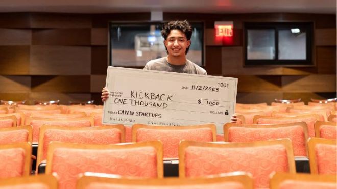 A student stands in the middle of an empty row of seats holding a large $1,000 check.