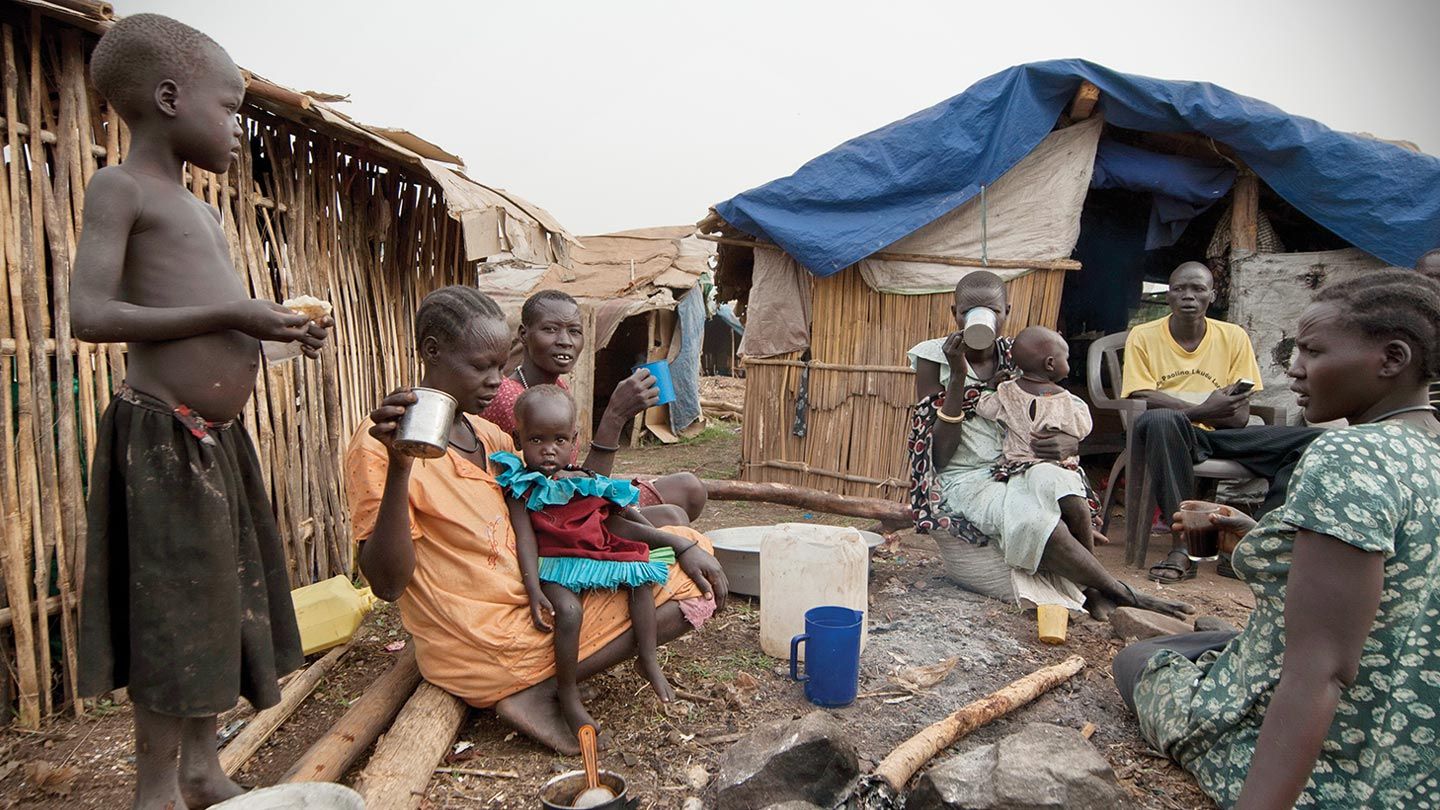 People in South Sudan with little resources sit together by an area with wood huts.