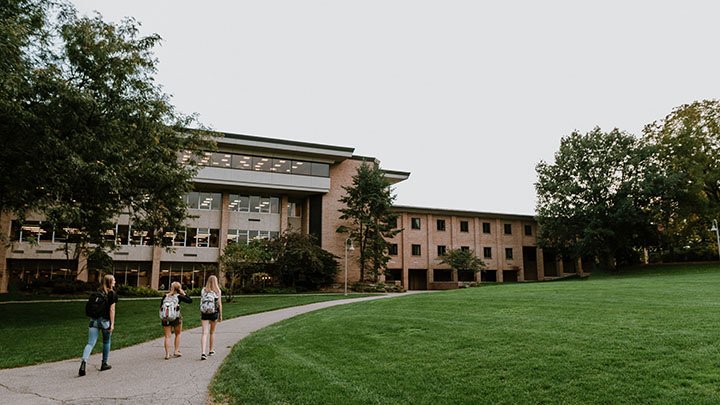 Three students walk away from the camera past a wide expanse of green space toward a campus building