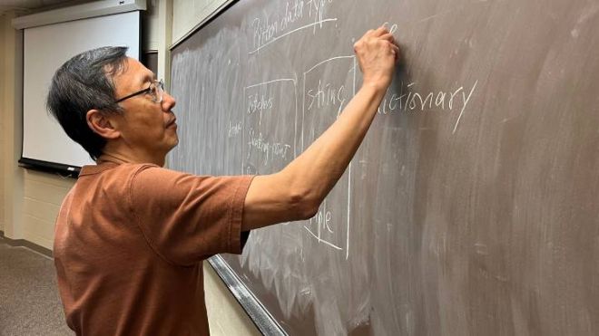 A man in a brown shirt and glasses writes on a chalkboard.