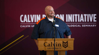A man wearning prison blues smiles at a podium with a Calvin Prison Initiative banner behind him.