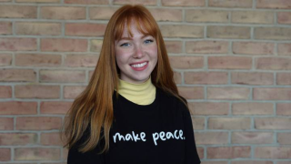 A female student smiles at the camera with a "make peace" shirt on.