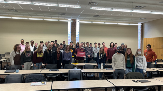A classroom of students poses with a professor holding a sign saying "Professor of the Year"