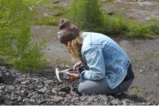 Jillian sitting with a pickaxe, observing a rock while doing research