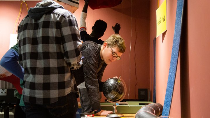 Three students examine a globe while a fourth student searches behind a lifted poster on the wall.