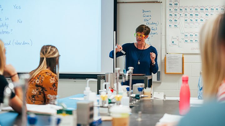 A professor wearing goggles performs an experiment in a lab with students looking on.