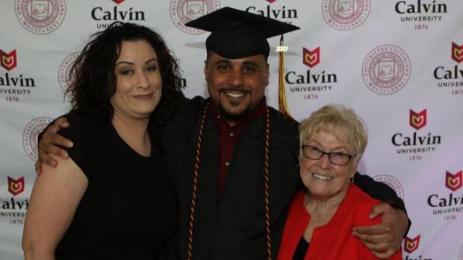A man with a cap and gown embraces family in front of a Calvin University backdrop.