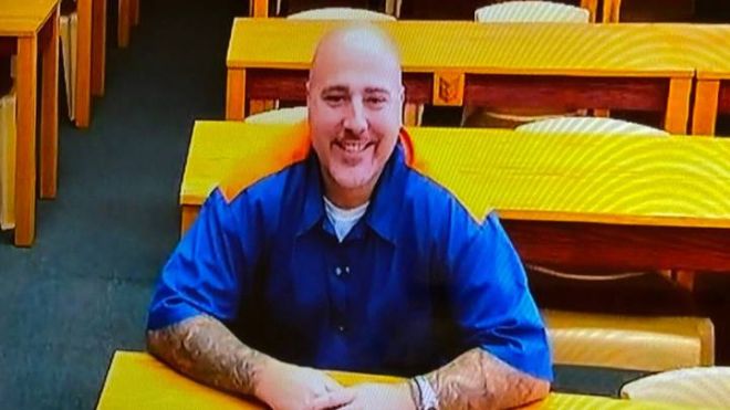 A man in prison blues smiling seated at a desk.
