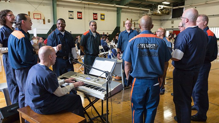 A group of prisoners stand in a circle, one playing the piano, the rest singing.