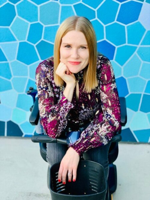 Amy, a white woman with shoulder-length, straight, blonde hair, sits in a mobility scooter wearing a burgundy blouse and grey jeans, in front of a wall with blue geometric shapes.
