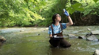 Female student wading in Plaster Creek, examining a sample of water, holding it up to the sky.