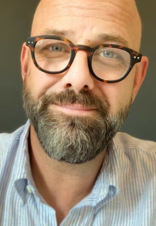 Gregory, a white man with a brown beard, bald head and tortoise shell glasses poses for a close-up headshot wearing a blue and white button-up shirt.