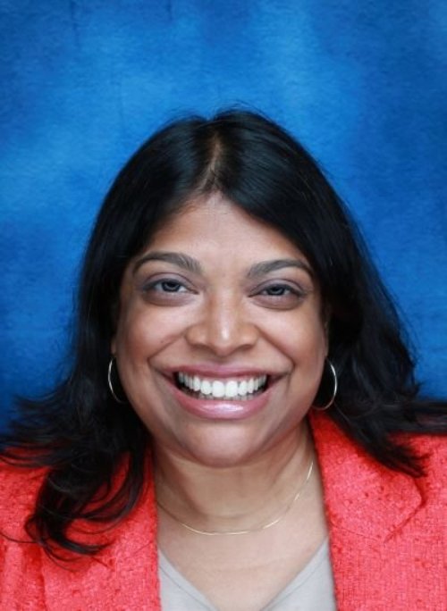Niala, an Asian American woman with long black hair, and silver hoop earrings, wears a bright pink suit jacket in a professional headshot.