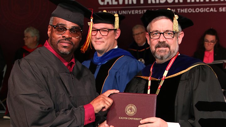 An African American male wearing glasses holds his diploma and poses with a white male president.