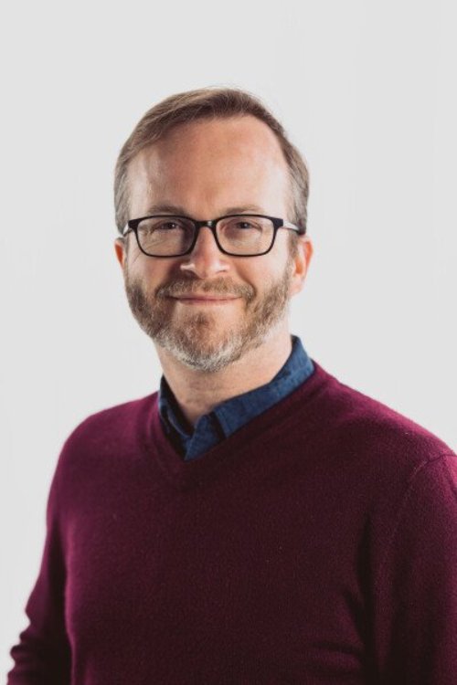 Tim, a white man with sandy brown hair and beard with glasses, wears a maroon sweater with a blue-collar shirt in a professional headshot with white background.