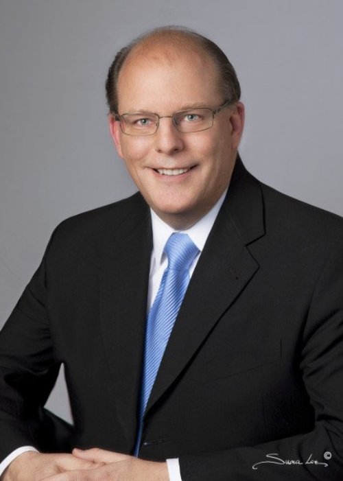 Peter, a white man with brown hair and glasses posts for a head shot in front of a grey background wearing a black suit coat, white button-up shirt and light blue tie.
