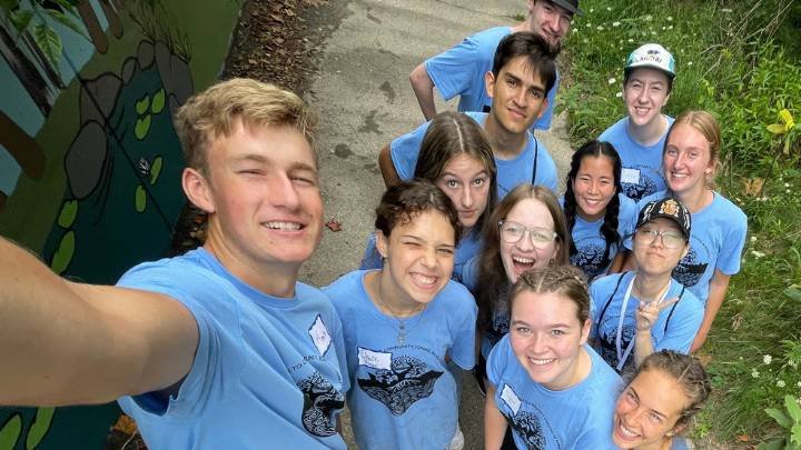 A group of 12 students wearing light blue T-shirts pose together for a selfie photo.