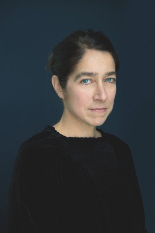 Kerri, a white woman with brown hair tied back poses for a head shot in a black shirt in front of a dark blue background.