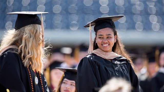 Two graduates in caps and gowns stand and smile at one another.