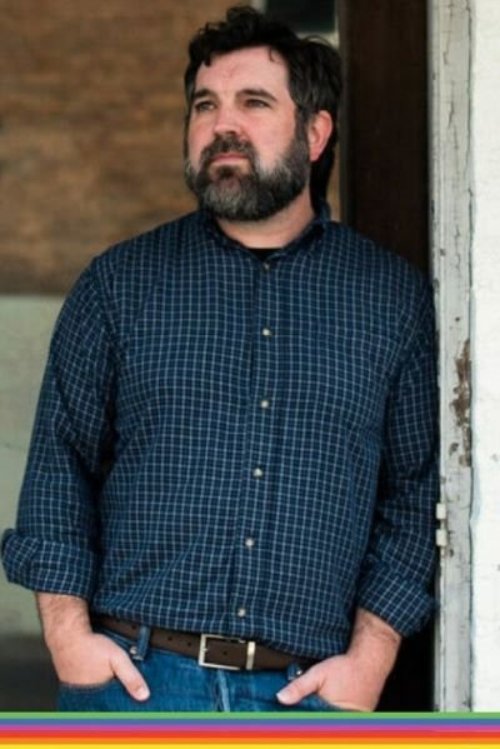 Daniel, a white man with black hair and a black beard, stands leaning against a pole wearing a blue patterned button up shirt and jeans.