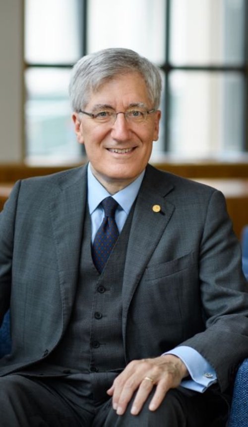 Robert, a white man with grey hair and glasses, sits in a chair wearing a dark grey suit, in front of windows.