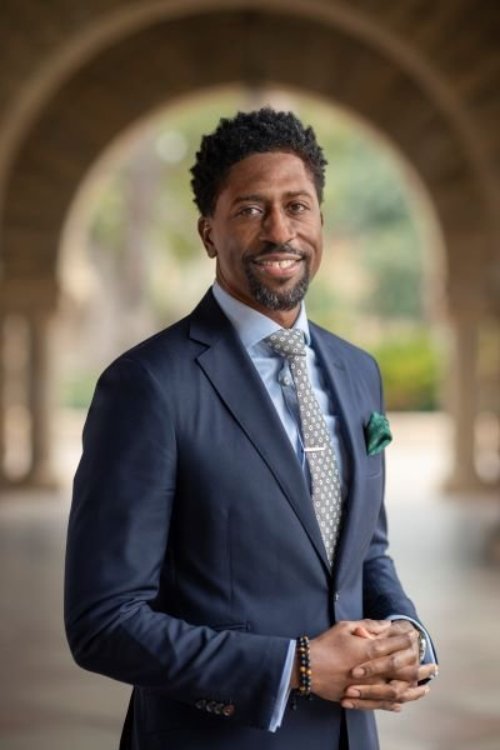 Lerone, a Black man with a goatee beard, stands wearing a navy-blue suit in front of an archway outside.
