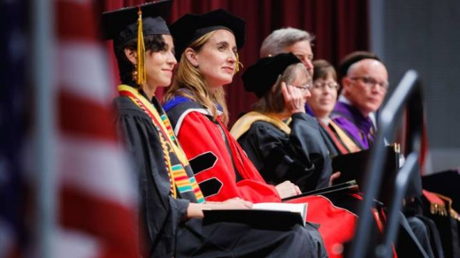 A woman dressed in red academic regalia is seated on stage alongside the platform party.