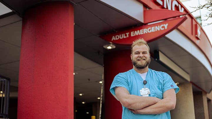 A student in short-sleeved blue scrubs with arms folded in front of hospital's emergency entrance.