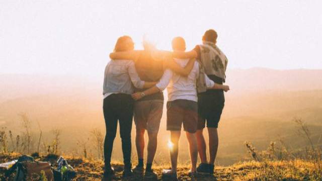 Four young adults look out at sunset over mountains, backs turned to camera, arms around each other