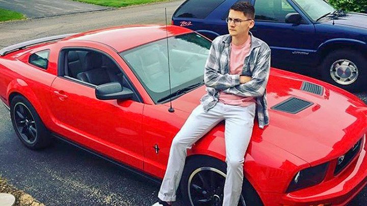 A young man sits on a red mustang car.