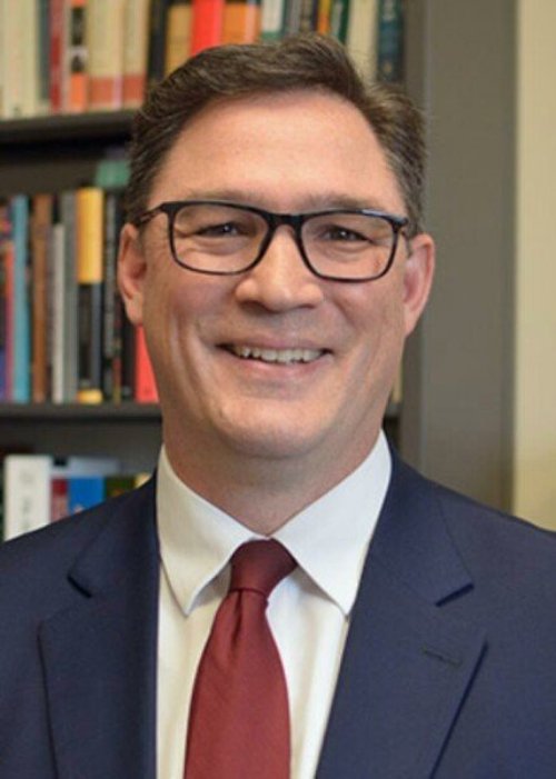 Noah, a white man with brown hair and glasses poses for a head shot in front of a bookcase wearing a blue suit coat, white button-up shirt and red tie.