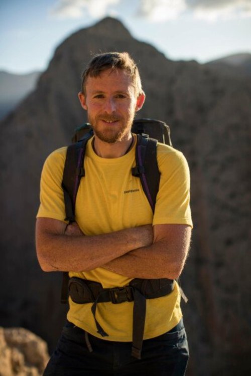 Tommy, a white man, with sandy brown hair, stands wearing a yellow t-shirt and hiking backpack in front of mountains.