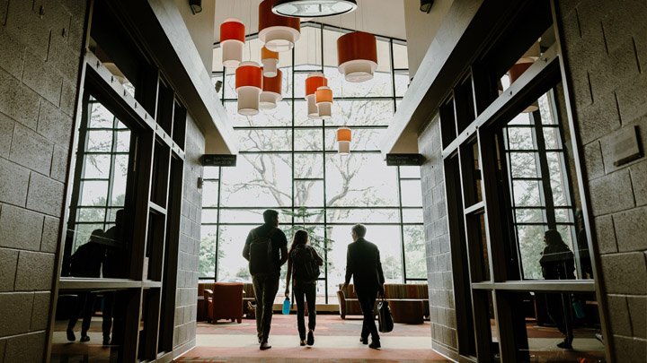 Two students and a professor talking while exiting a hallway are silhouetted against a large window.