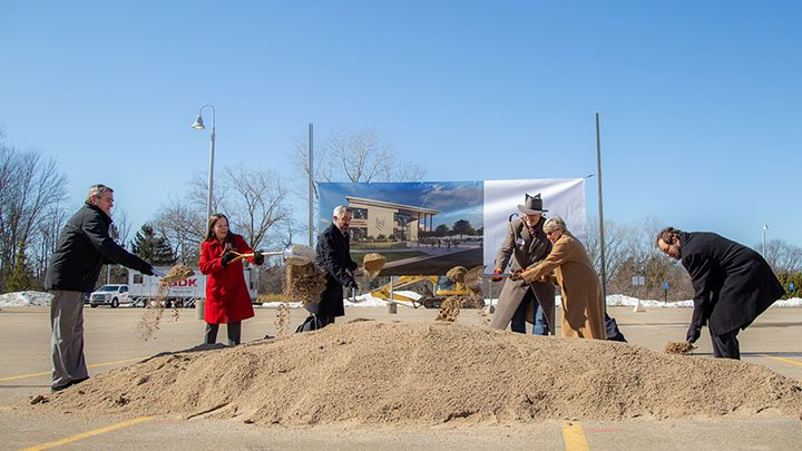 Six people with shovels in hand scoop up dirt in front of a rendering of the new building.
