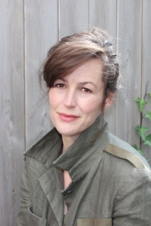 Sara, a white woman with brown hair tied back, poses for a headshot wearing an army green blouse, in front of a fence.