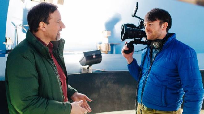 A man in a green jacket talks to a man in a blue jacket holding a camera inside an observatory.