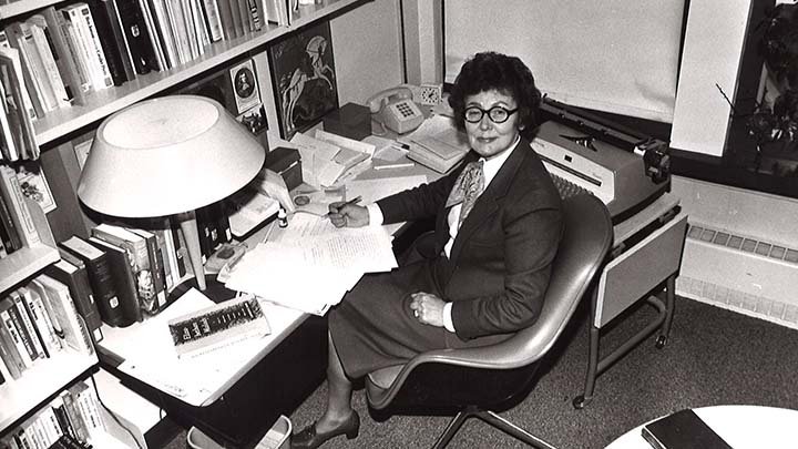 A black and white photo of a woman in a suit jacket and a skirt sitting at a desk near a bookshelf.