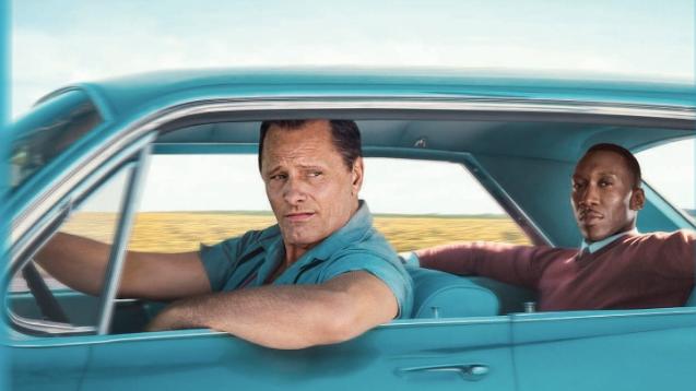 Two main characters sit in a car--one man driving the other. Driver has his arm out the window.