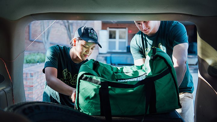 Two college students load a bag of uneaten food into the trunk of a car.