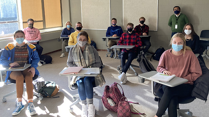 Class of high school students in masks sit in desks with college professor standing in back of room