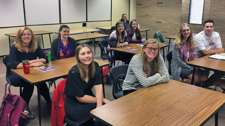 Nine smiling students sit at shared tables facing forward in a classroom.
