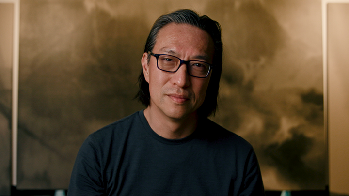 A man in glasses and a black shirt looks intently straight ahead with artwork behind him.