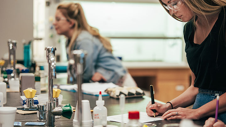 Two female students stand by and do work at a lab table.