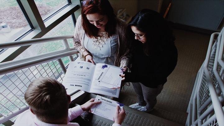 Three students are standing in a stair well talking while looking at papers they are holding