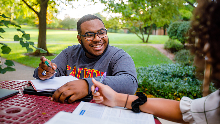 On a fall day,  a student smiles as he sits at a table outside across from two fellow students.