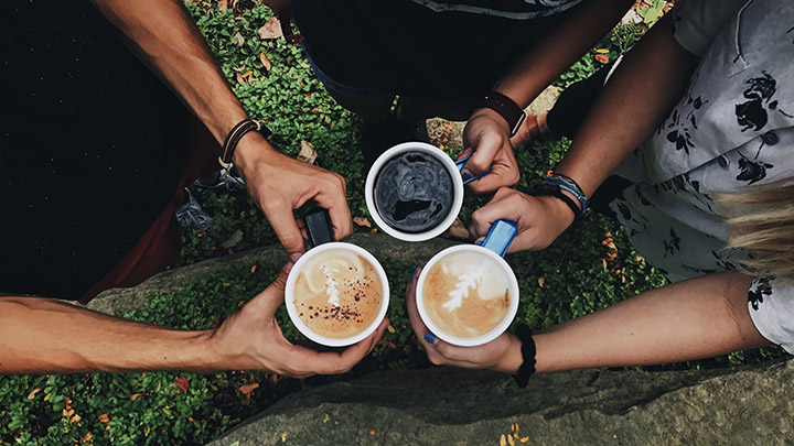 Three people hold full coffee mugs together in triangle above grass.