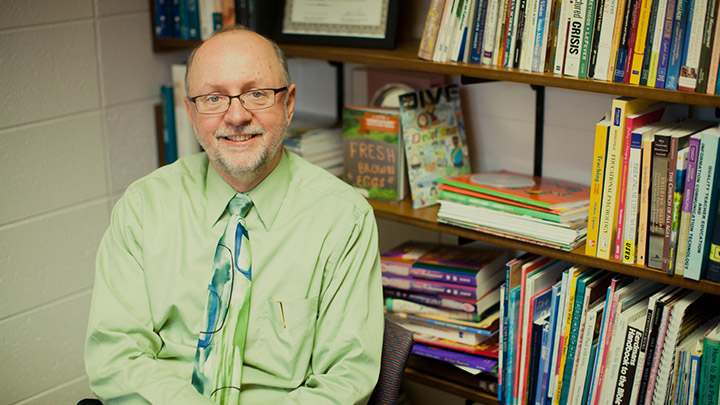 A male professor in a shirt and tie smiles sitting in front of his office bookshelf.