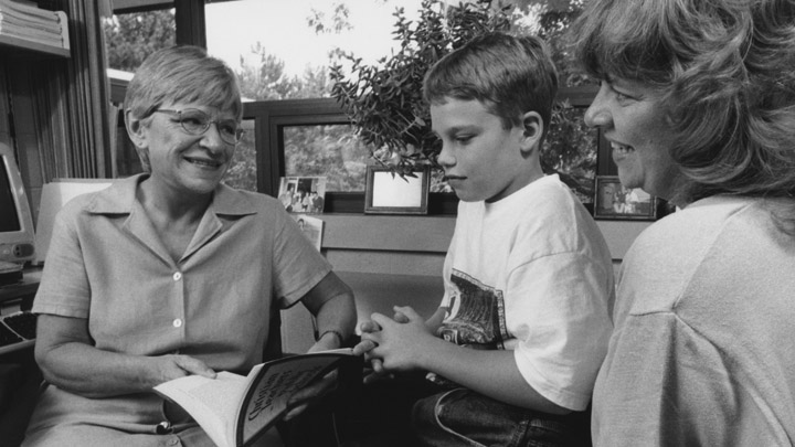 A middle-aged woman holding a book sitting and talking with a child and his mother.