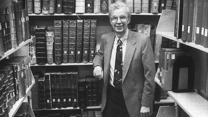 A man in a suit coat and tie stands in a room with bookshelves on three sides.