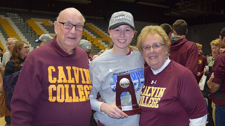 Two grandparents pose with their granddaughter holding a national championship volleyball trophy.